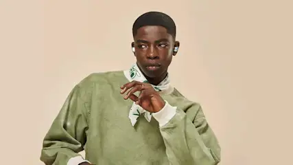 Man met oortjes draagt een olijfgroene trui en een witte sjaal met groene motieven tegen een beige achtergrond.