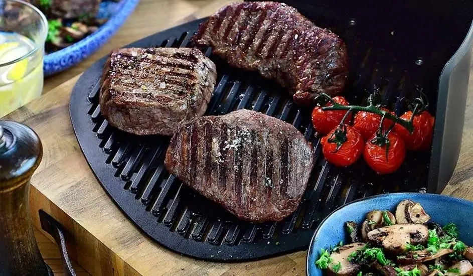 On a Tefal contact grill lie three pieces of grilled meat and a cluster of tomatoes. Next to the grill stands a plate with fried mushrooms.