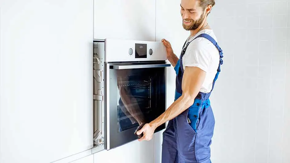 Een man in blauwe overall installeert een witte oven in een witte kast in een witte kamer.