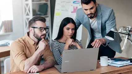 Drie personen werken aan een bureau met een grijze laptop. Er staat een witte mok en papieren op het bureau.