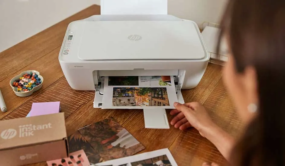 Woman using a white HP printer on wooden table, colourful photos sliding out beside Instant Ink box and craft supplies.