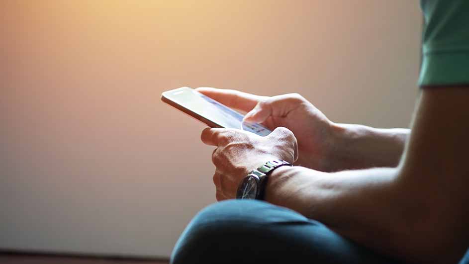 Een persoon zit met een telefoon in de hand. De persoon draagt een horloge en een groen shirt. De achtergrond is effen.