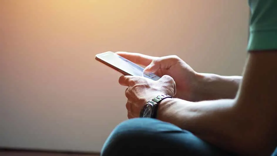 Een persoon zit met een telefoon in de hand. De persoon draagt een horloge en een groen shirt. De achtergrond is effen.