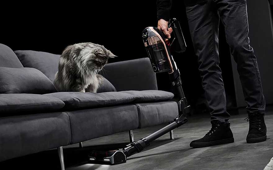 Een man stofzuigt rond een kat op de bank met een steelstofzuiger, krachtig en handig voor huisdierenbezitters.
