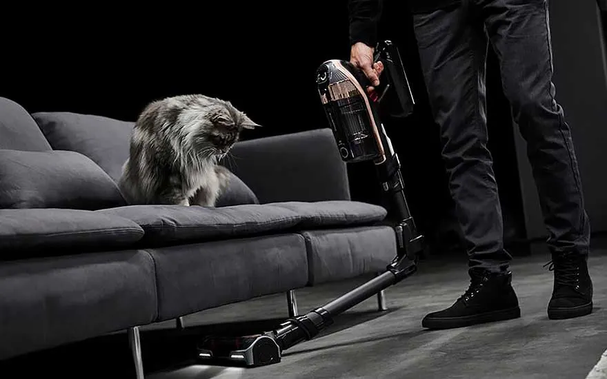 Een man stofzuigt rond een kat op de bank met een steelstofzuiger, krachtig en handig voor huisdierenbezitters.