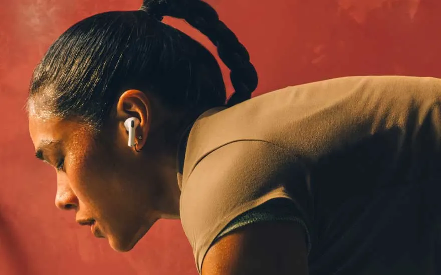 A person in sportswear leans forward, sweating and concentrated, wearing white wireless earbuds against a warm orange-coloured background.
