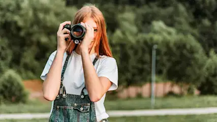 Een persoon in een wit shirt en blauwe tuinbroek houdt een camera vast in een groene, buiten omgeving.