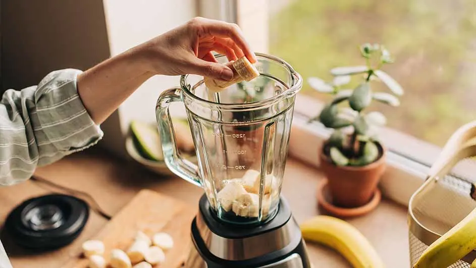 Hand voegt banaan toe aan een blender op een houten aanrecht. Bananen, snijplank, plant en raam zijn zichtbaar.