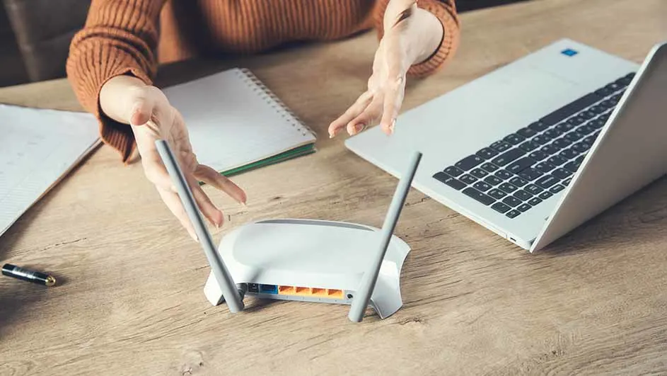 Op een houten bureau staan een witte router, een laptop, notitieblokken en een pen, met handen erboven.