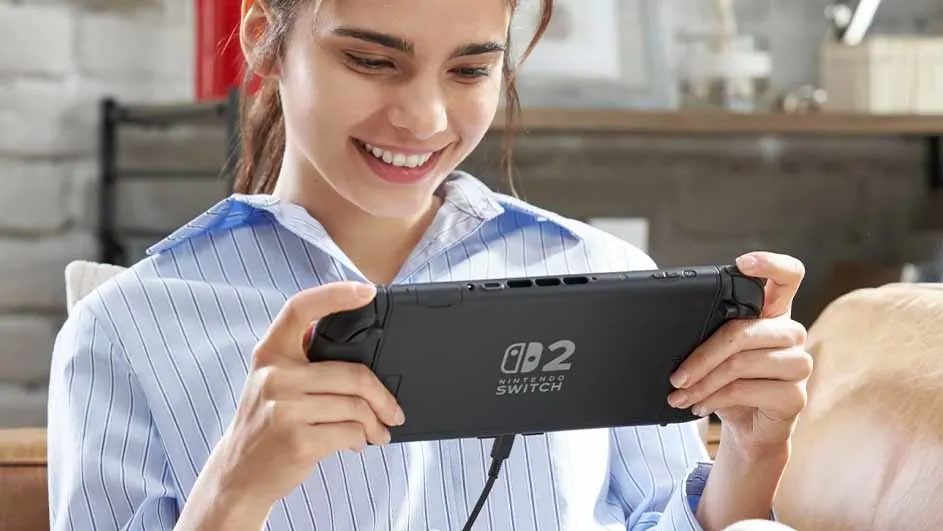 Een lachende persoon houdt een zwarte Nintendo Switch vast, met een kabel eraan bevestigd. De persoon draagt een blauw gestreept overhemd.