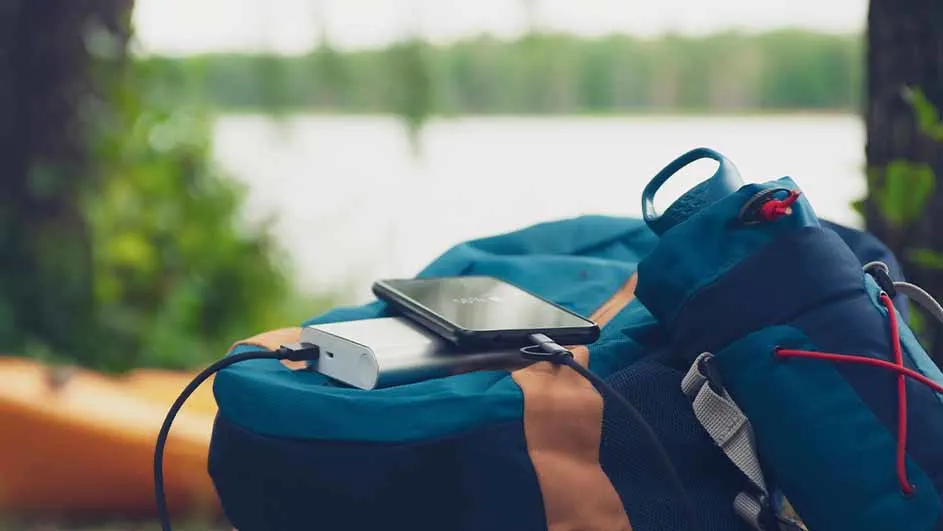 Draagbare powerbank die een smartphone oplaadt op een blauw met oranje rugzak in een bosrijke omgeving bij een meer.