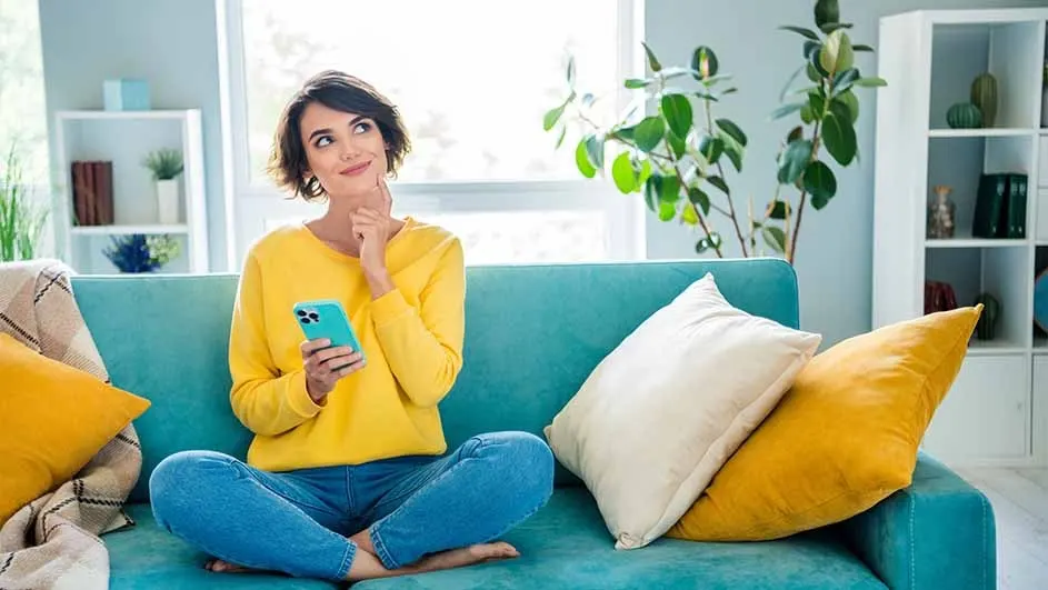 Person sits cross-legged on a blue sofa with pillows, holding a blue phone and looking thoughtful in a bright room.
