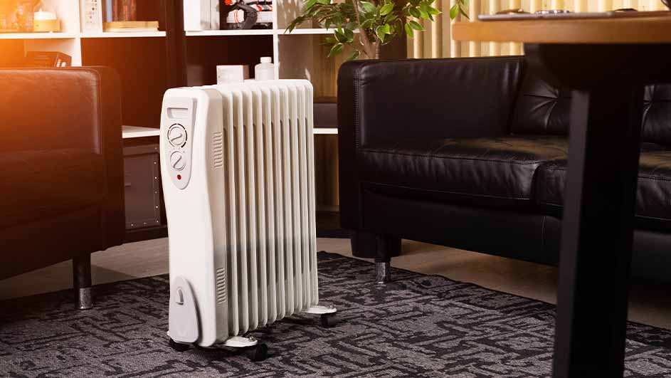 Een kamer toont een witte oliegevulde radiator op een grijze mat, met een zwart leren bank en houten meubels op de achtergrond.