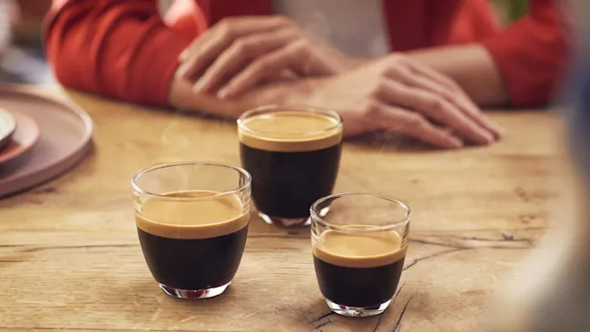 Drie glazen espresso op een houten tafel, met op de achtergrond een persoon in een rood vest.