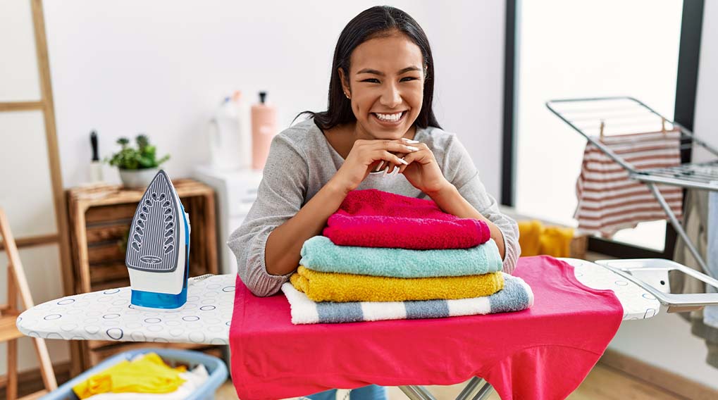 Een vrouw glimlacht achter een stapel handdoeken op een strijkplank, met een strijkijzer ernaast. Op de achtergrond staat een droogrek.