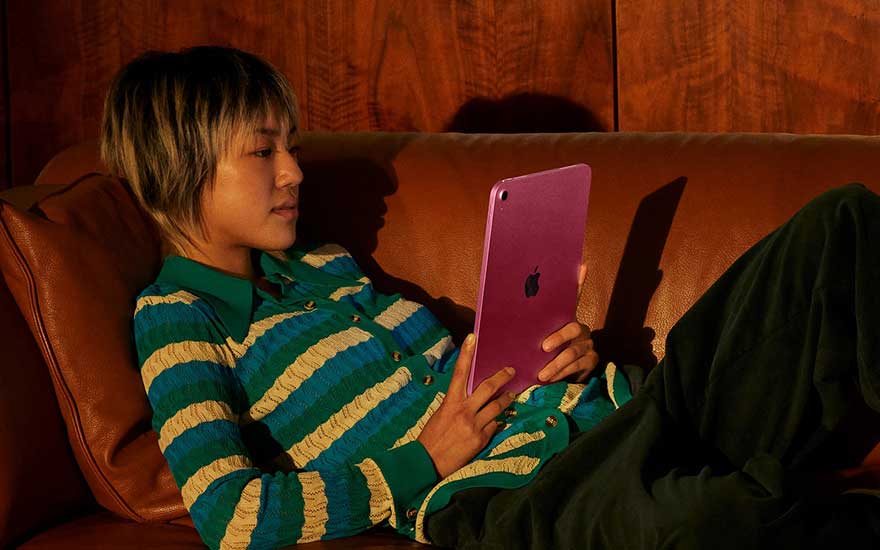 Een persoon ligt ontspannen op een leren bank en houdt een paarse tablet vast; warm licht benadrukt het houten paneel en de gezellige sfeer.