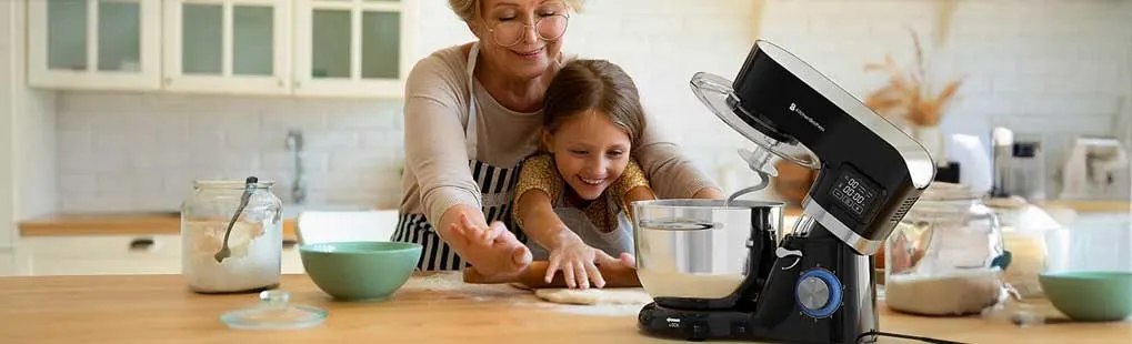 Oma helpt haar kleindochter deeg rollen op een houten aanrecht, terwijl een zwarte keukenrobot beslag mengt in een glimmende kom.