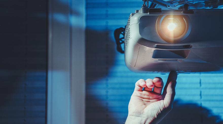 Een hand raakt een grijze projector aan die aan het plafond hangt, met een blauwe achtergrond.