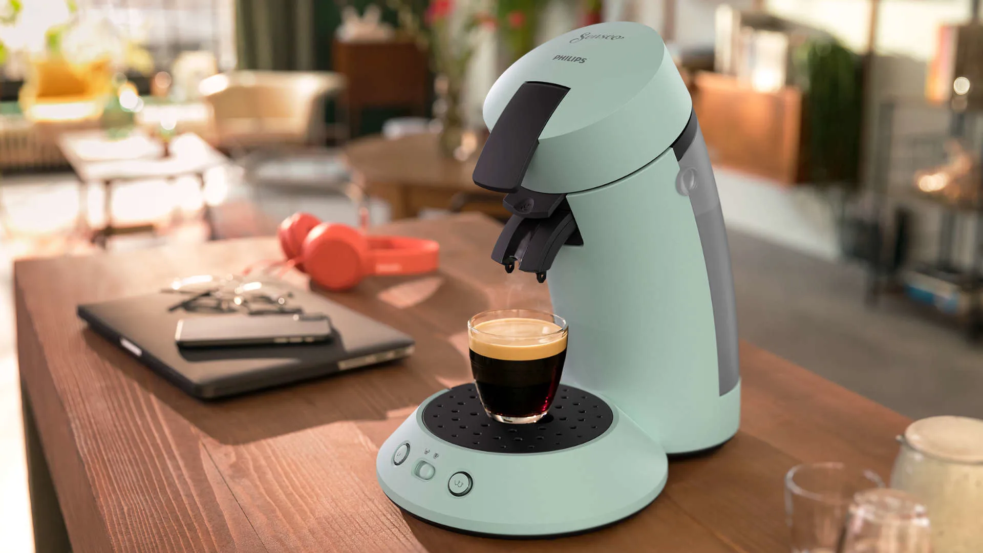 Een mintgroen Senseo koffiezetapparaat met een kopje koffie op een houten tafel, met laptop en koptelefoon op de achtergrond.