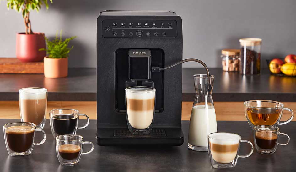 Een zwarte Krups koffiemachine staat op het aanrecht, omringd door acht glazen met verschillende koffievariaties, melk en thee.
