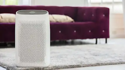 Witte luchtreiniger op een grijze vloerkleed, met een paarse sofa op de achtergrond.