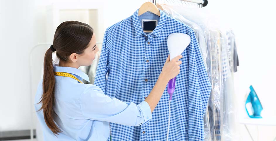 Een vrouw stoomt een blauw geruit overhemd aan een houten hanger, met kledingrekken en een blauw strijkijzer op de achtergrond.