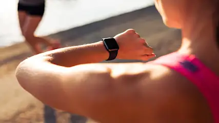 Een vrouw draagt een zwarte smartwatch en een roze top, met een vage figuur in de achtergrond op een zandkleurige ondergrond.