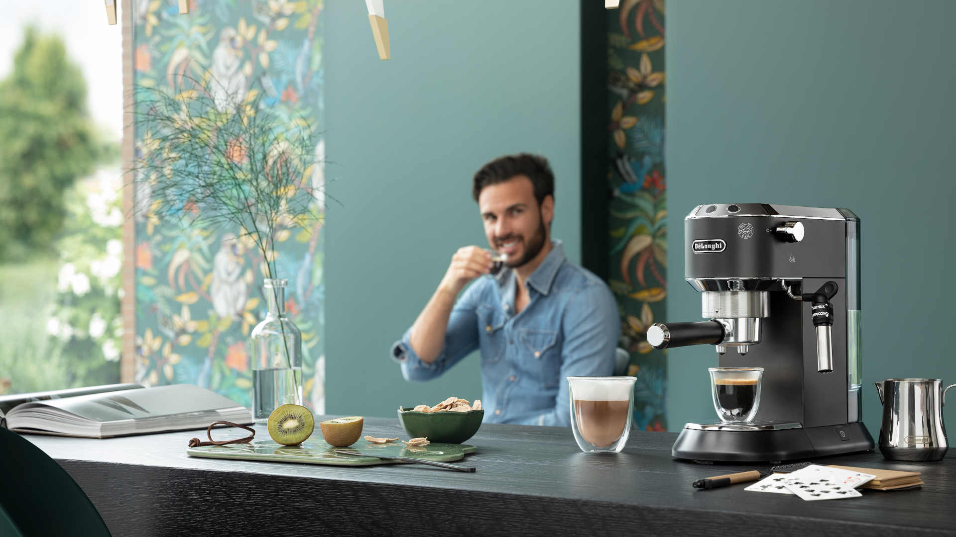 Een zwarte DeLonghi espressomachine staat op een tafel met een kopje koffie, melkkan en notities. Op de achtergrond zit een man koffie te drinken.