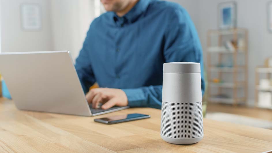Een man typt op een laptop aan een houten tafel met een telefoon en een witte speaker. Op de achtergrond is een kamer te zien.