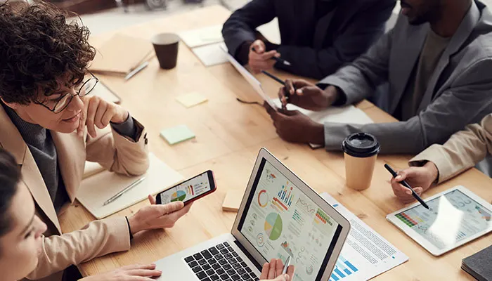Team op kantoor zit rond een tafel met laptop, tablet, telefoon en koffiebekers. Grafieken zijn zichtbaar.