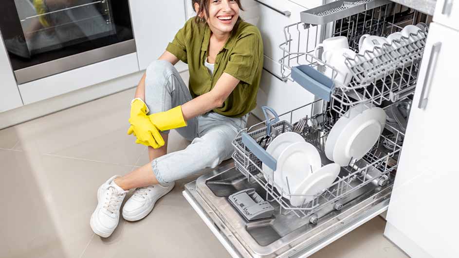 Vrouw met gele handschoenen zit op de grond naast een open vaatwasser vol witte borden en kopjes in een keuken.