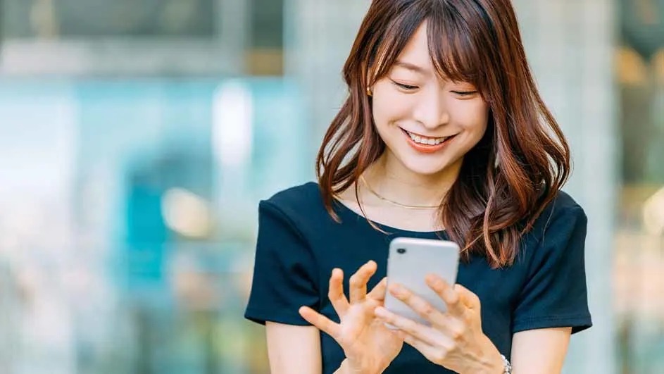 A smiling woman in a navy top looks down at her phone, fingers tapping the screen as soft daylight blurs the busy city background.