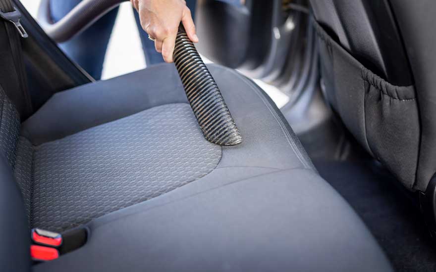 Close-up of a hand using a vacuum cleaner hose to thoroughly clean the back seat of a car, removing visible dust and dirt.