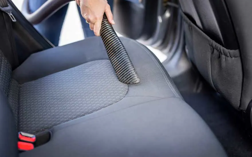 Close-up of a hand using a vacuum cleaner hose to thoroughly clean the back seat of a car, removing visible dust and dirt.