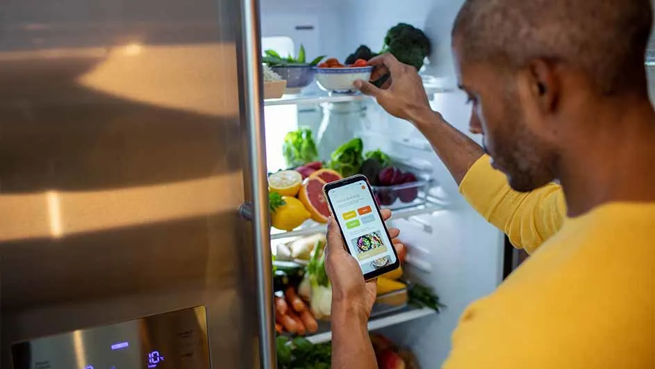 Een man in een geel shirt kijkt in een koelkast vol groenten en fruit, terwijl hij een telefoon vasthoudt.