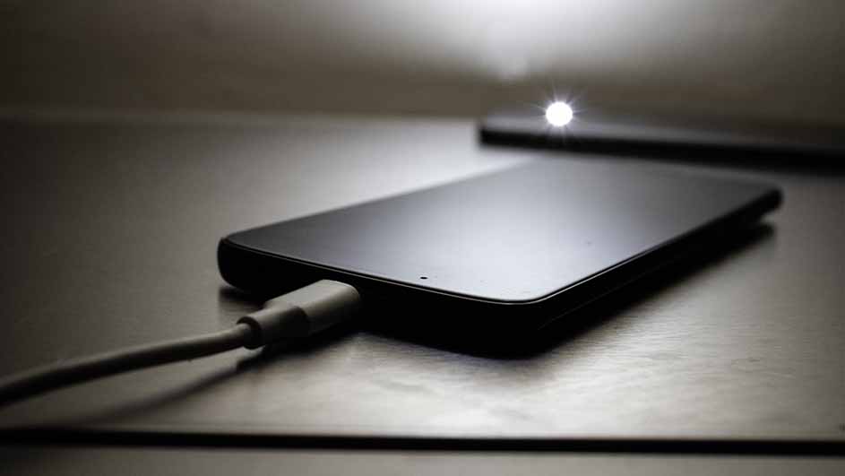 Een zwarte smartphone ligt op een tafel en wordt opgeladen met een grijze USB-kabel. Een ander apparaat ligt op de achtergrond.