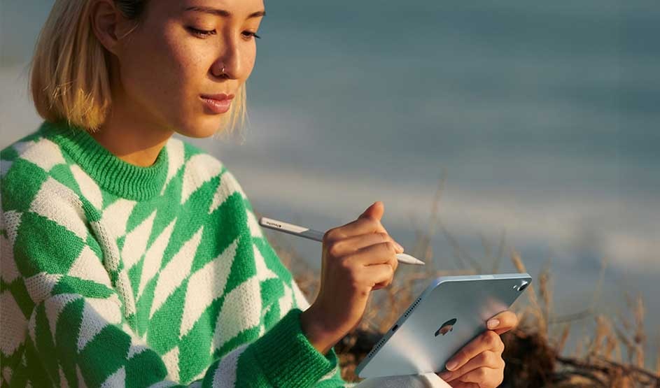 Woman sitting outside by the sea, drawing intently on a small blue tablet with stylus, wearing a green jumper with white pattern.