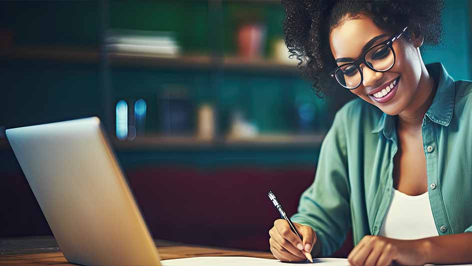 Een vrouw met een bril schrijft aan een tafel met een pen op papier, naast een laptop. Achtergrond: een vage groene plank.