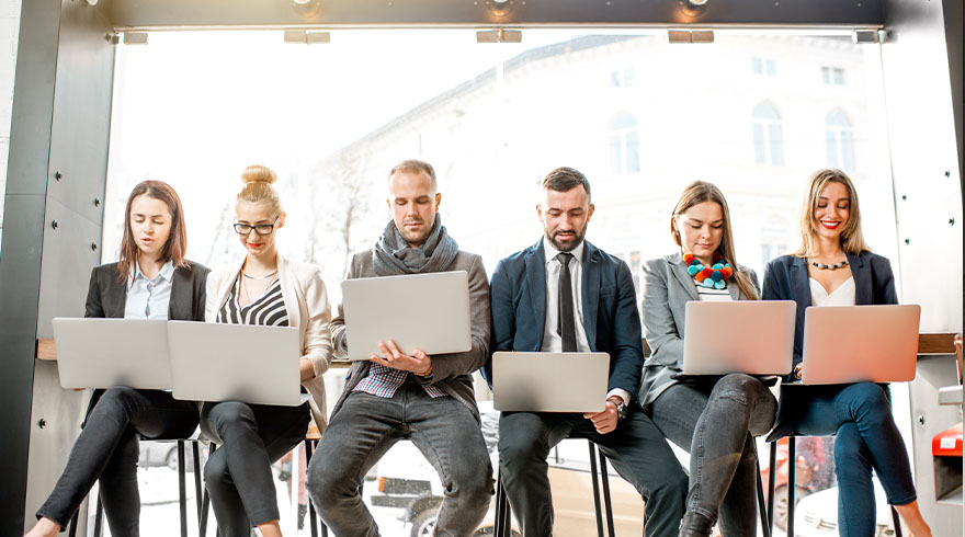 Vijf mensen zitten op stoelen met laptops. Allen dragen formele of semi-formele kleding. Een raam is zichtbaar op de achtergrond.