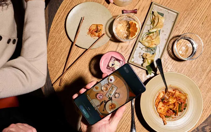 Bovenaanzicht van een houten tafel met Aziatische gerechten, drankjes en stokjes, terwijl iemand met een Samsung smartphone een foto van het eten maakt.