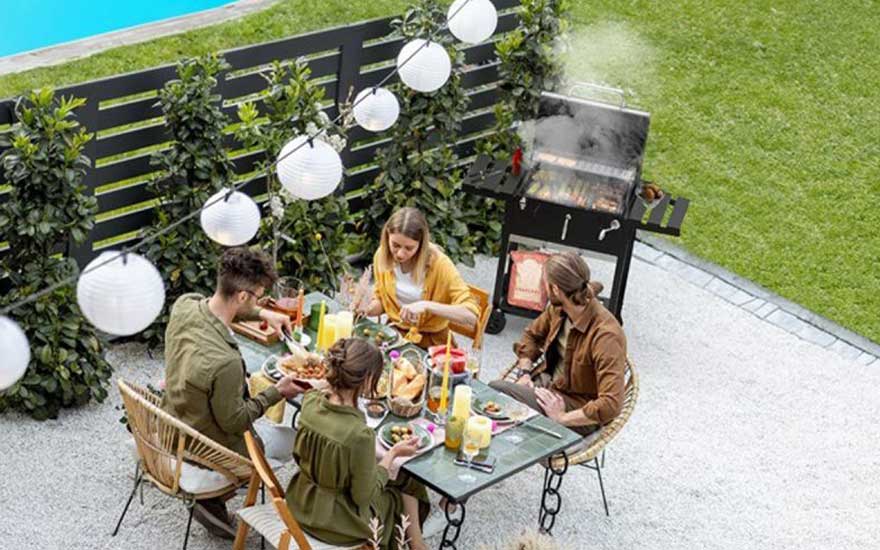Four friends share a meal on a tidy patio, with lanterns, colourful drinks and a barbecue smoking in the background.