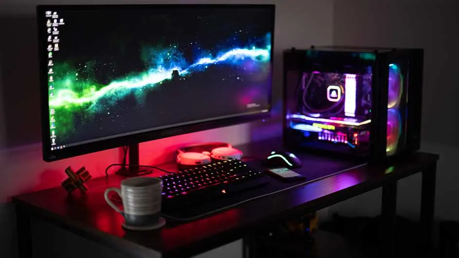 Een bureau met een computer, toetsenbord, muis, koptelefoon, mok en een decoratief object. De computer heeft kleurrijke verlichting.