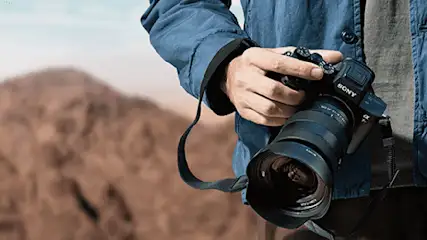Een persoon houdt een zwarte Sony camera vast met een grote lens. Op de achtergrond is een bruine heuvel te zien.