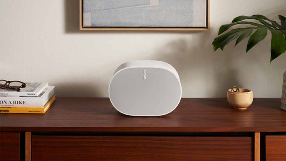 Een witte Sonos speaker staat op een houten dressoir naast boeken, een plant en een houten schaaltje.