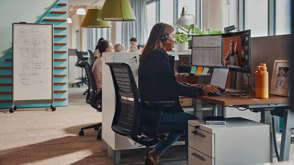 Een vrouw met headset werkt aan een bureau met een laptop en monitor in een kantoorruimte. Een whiteboard staat op de achtergrond.