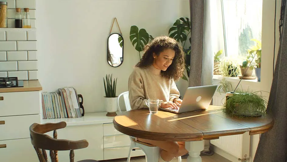 Vrouw typt op een laptop aan een ronde houten tafel. Planten en een spiegel staan op de witte kast. Een raam zit achter haar.