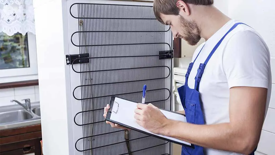 Een man in blauwe overall inspecteert de achterkant van een witte koelkast met een zwart raster en maakt aantekeningen op een klembord.