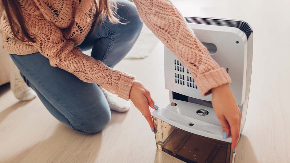 Een persoon knielt en leegt de waterbak van een witte luchtontvochtiger op een houten vloer.