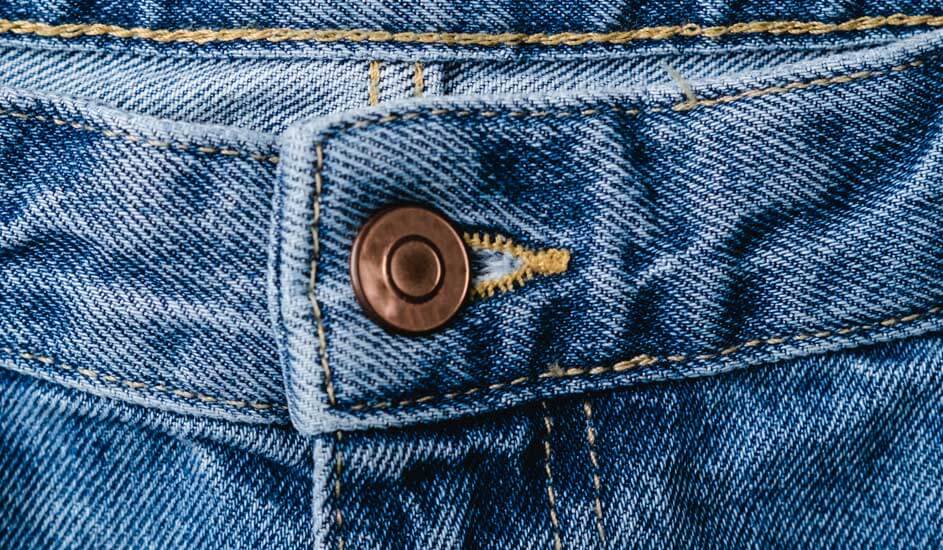 Een close-up van een blauwe spijkerbroek, waarop de tailleband, een metalen knoop en gele stiksels duidelijk zichtbaar zijn.