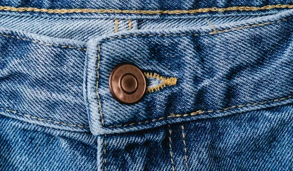 Een close-up van een blauwe spijkerbroek, waarop de tailleband, een metalen knoop en gele stiksels duidelijk zichtbaar zijn.